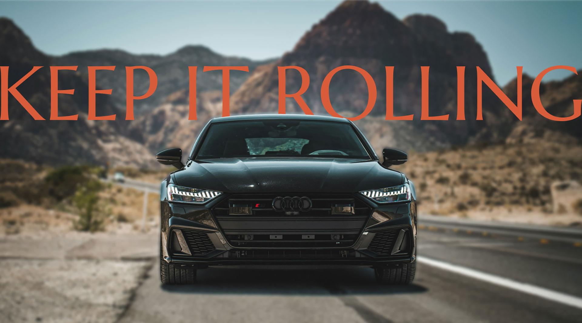 KeepItRolling Front view of a sleek black Audi driving on a desert highway with mountains in the background. Bold orange text across the top reads ‘KEEP IT ROLLING.