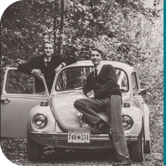 about-us-secondary-img “Black-and-white photo of two men posing with a classic Volkswagen Beetle on a wooded road. One man leans against the open driver’s door, while the other sits on the car’s front fender.