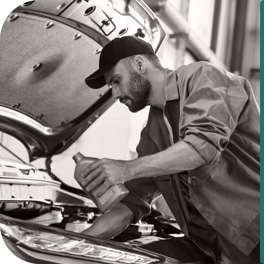 Black-and-white photo of an auto mechanic in a cap and uniform adding motor oil to a car engine with the hood open in a service garage.
